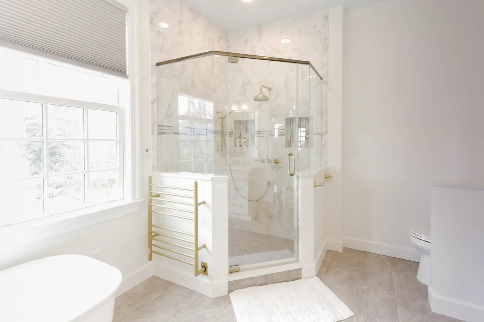 Bright and elegant bathroom featuring a frameless swinging glass enclosure with gold hardware, marble walls, and a luxurious spa-like ambiance.